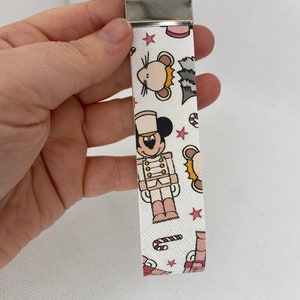 May include: White keychain with a pink and brown pattern featuring Mickey Mouse dressed as a nutcracker, a mouse, candy canes, and stars.