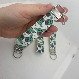 May include: White keychain with a repeating pattern of green cartoon characters. The keychain has a silver metal ring and a silver metal loop.