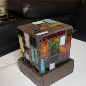 May include: A square lamp featuring a stained glass shade constructed from various rectangular glass pieces in red, orange, yellow, green, blue, white, and black colors. The shade is mounted on a dark brown wooden base with a white power cord.