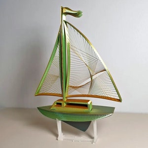 Peut inclure: Une sculpture de voilier verte et dorée avec une voile en treillis métallique. Le voilier est sur un support en plastique transparent.