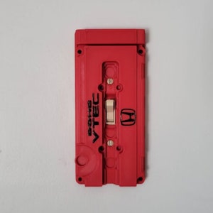 May include: A red plastic light switch cover with a black Honda logo and the words "VTEC" and "Synchro" printed on it. The switch is white and is in the off position.
