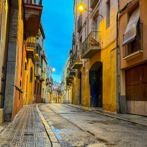 May include: An urban street scene at dusk. Buildings with colorful facades line the cobblestone street, illuminated by streetlights. The sky is a deep blue, contrasting with the warm tones of the buildings.