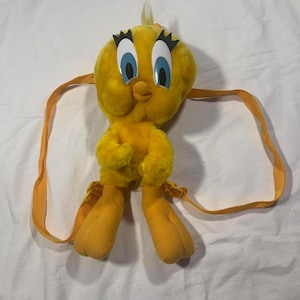 Może przedstawiać: Żółty pluszowy plecak Tweety Bird z pomarańczowymi szelkami. Plecak wykonany jest z miękkiego, puszystego materiału i ma charakterystyczne duże oczy i dziób Tweety'ego.