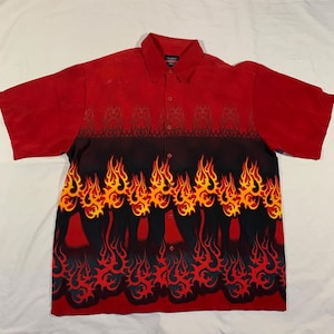 Puede incluir: Camisa roja de manga corta con botones y un patrón de llamas. Las llamas son amarillas, naranjas y rojas, y están sobre un fondo negro. La camisa tiene un cuello abotonado y un frente abotonado.