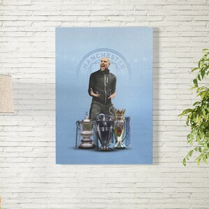 May include: A man in a black jacket and jeans stands in front of a blue background with the word "Manchester" in a circle. He is holding up a trophy with a silver base and a gold top. There are two other trophies behind him.