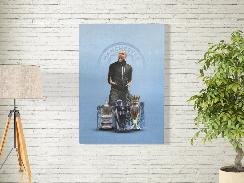 May include: A man in a black jacket and jeans stands in front of a blue background with the word "Manchester" in a circle. He is holding up a trophy with a silver base and a gold top. There are two other trophies behind him.