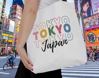May include: A white tote bag with the words "TOKYO TOKYO Japan" printed in colorful, overlapping letters. The bag is being held by a person in front of a busy street in Tokyo, Japan.