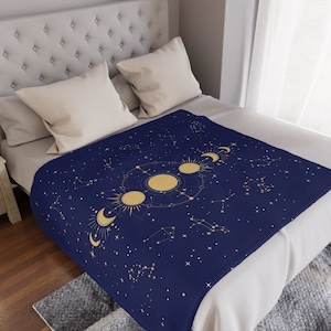 May include: A navy blue throw blanket with a gold celestial design featuring constellations, moons, and stars. The blanket is on a bed with white bedding.