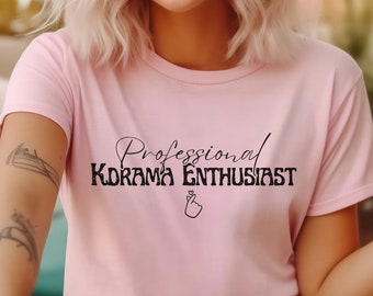 Puede incluir: Una camiseta rosa con el texto "Professional Kdrama Enthusiast" en letras negras. El texto está en una fuente cursiva e incluye un pequeño corazón y un gesto de mano.