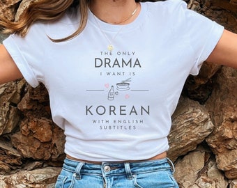 Puede incluir: Camiseta blanca con el texto "The only drama I want is Korean with English subtitles". El texto está rodeado por una línea y corazones. Hay un dibujo de un tazón de fideos, palillos y una botella de soju.