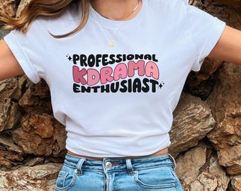 Puede incluir: Camiseta blanca con el texto "PROFESSIONAL KDRAMA ENTHUSIAST" en letras de burbuja negras y rosas.