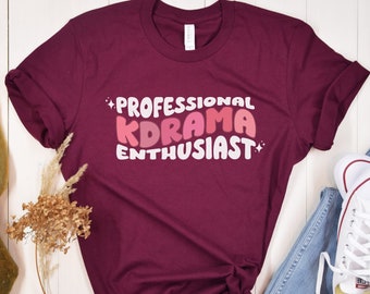 Puede incluir: Una camiseta color burdeos con el texto "Professional Kdrama Enthusiast" en letras rosa, blancas y rojas.