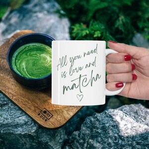 Pode incluir: Caneca de cer&acirc;mica branca com o texto "All you need is love and matcha" em escrita verde com um pequeno cora&ccedil;&atilde;o desenhado abaixo. Uma m&atilde;o est&aacute; segurando a al&ccedil;a da caneca. Uma tigela de ch&aacute; matcha verde est&aacute; no fundo.