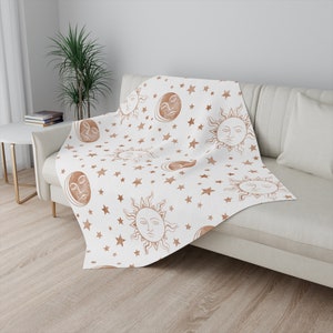 May include: A white throw blanket with a repeating pattern of suns, moons, and stars in a brown watercolor design.