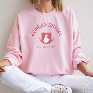 May include: A pink sweatshirt with the text "Korean Drama University" in a pink and white font. A pink heart and a hand gesture are inside a shield with a laurel wreath.