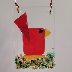 May include: A rectangular glass ornament featuring a red bird design. The bird has a yellow beak and a black eye. The bird is perched on a bed of colorful glass pieces. Copper wire is used for hanging.