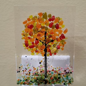 May include: A decorative night light featuring a fused glass design of a tree with a black trunk and a colorful mosaic of orange, yellow, and red glass pieces for the leaves. The base of the tree has a colorful mosaic of glass pieces. The night light plugs into a standard wall outlet.
