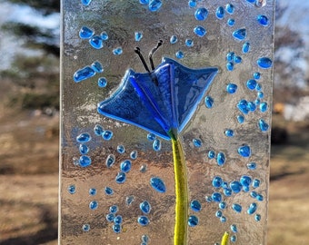 May include: A blue glass flower with a green stem and leaves, surrounded by small blue, green, and yellow glass beads. The flower is hanging from a copper wire.