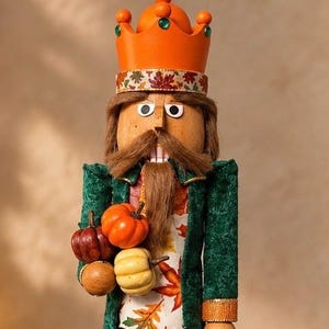 May include: A wooden nutcracker figure wearing an orange crown with green accents and a green velvet coat. The figure has a brown mustache and beard, and holds a cluster of small orange, yellow, and dark red pumpkins. The vest features a colorful autumn leaf pattern.