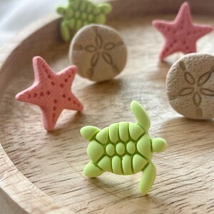 以下が含まれることがあります： ピンクのヒトデ、緑のカメ、砂色のサンドダラーなど、海のテーマのミニチュアフィギュアのコレクション。 フィギュアは丸い明るい色の木製トレイに配置されており、海岸の美学を作り出しています。