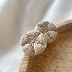 以下が含まれることがあります： ベージュ色の砂ドル型のスタッドピアス2つ。ピアスは、テクスチャ加工された表面と、詳細で対称的なデザインが特徴です。木製のトレイの上に、白い布の背景で表示されています。