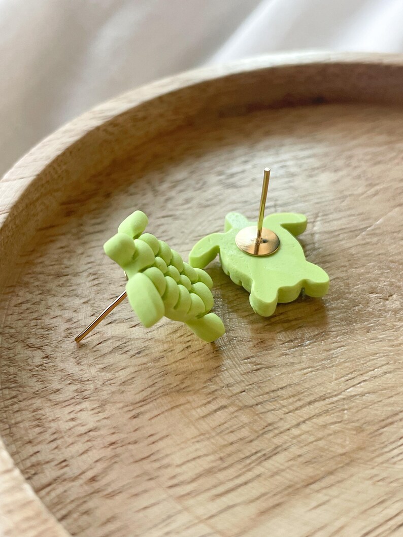 以下が含まれることがあります： 明るい緑色のカメの形をしたスタッドピアス。片方はテクスチャ加工された甲羅を見せ、もう片方はポストと裏側を見せています。ピアスは木製の表面に置かれています。ピアスは明るい緑色の素材で作られています。