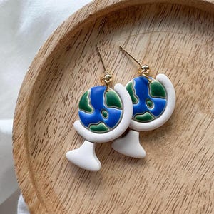 May include: A pair of globe-shaped earrings. Each earring features a blue and green globe design with white accents and gold-colored ear wires. The earrings are displayed on a wooden tray.