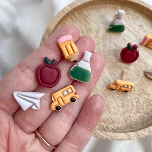 May include: A collection of colorful school-themed earrings. The earrings include a red apple, a yellow school bus, a pencil, a paper airplane, and a flask. The earrings are on a wooden tray.