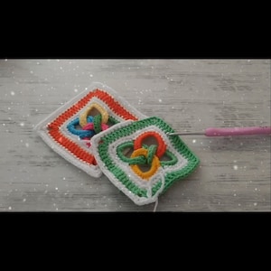 Peut inclure: Deux carrés au crochet avec un motif coloré et entrelacé. Les carrés sont faits de fil blanc, vert, orange, jaune et rose. Un crochet rose est visible sur l'image.