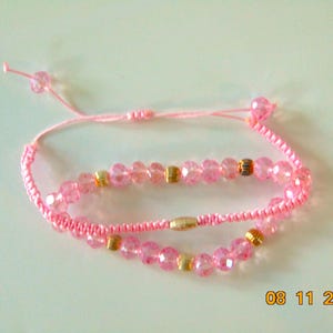 Könnte beinhalten: Ein rosafarbenes Kordelarmband mit facettierten rosa Perlen und kleinen goldfarbenen Metallzwischenstücken. Das Armband hat einen verstellbaren Schiebeknotenverschluss mit zwei baumelnden rosa Perlen an den Kordelenden, was einen Lagen-Look erzeugt.