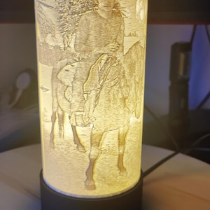 Pode incluir: Uma lâmpada cilíndrica com uma base preta e um abajur de vidro branco fosco. O abajur apresenta uma imagem gravada de uma pessoa a cavalo.
