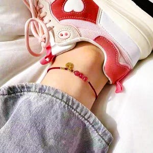 May include: A delicate anklet featuring a thin red cord, three small translucent red beads, and a circular gold-toned charm with an embossed character. The anklet is worn above a white and pink sneaker with a heart detail and blue denim fabric.