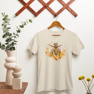 May include: A beige t-shirt featuring a watercolour illustration of a bee on a honeycomb.