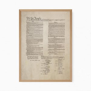 Könnte beinhalten: Ein gerahmter Druck der Verfassung der Vereinigten Staaten auf gealtertem, pergamentartigem Papier. Das Dokument zeigt die markante Überschrift We the People in schwarzer Tinte, gefolgt von Textspalten und zahlreichen Unterschriften, in einem hellen Holzrahmen.