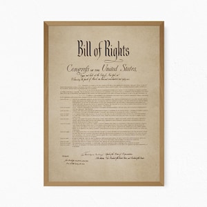 May include: A framed print of the Bill of Rights of the United States, featuring dark brown text on a light tan, parchment-style background. The document includes the title, preamble, and ten amendments, enclosed in a simple light brown wooden frame.