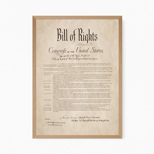 May include: A framed print of the Bill of Rights with a light brown wooden frame. The document features aged, parchment-style paper with black text, including the title Bill of Rights and Congress of the United States, displayed in a vertical orientation.