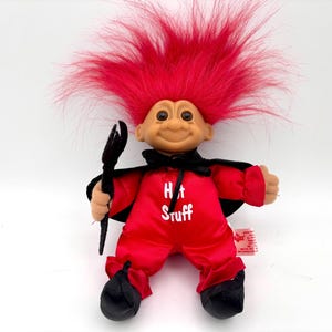 May include: A vintage troll doll with bright red hair and a smiling face. The doll wears a red satin outfit with the words "Hot Stuff" and a black cape. It holds a black trident.