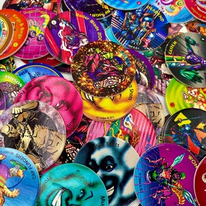 May include: A collection of circular cardboard game discs featuring colourful graphic illustrations of various characters, creatures, and text such as Mug Shots, Cap Flingers, and Glitch. The discs are layered in a pile, displaying diverse artistic styles, vibrant colours, and printed copyright dates from 1994.