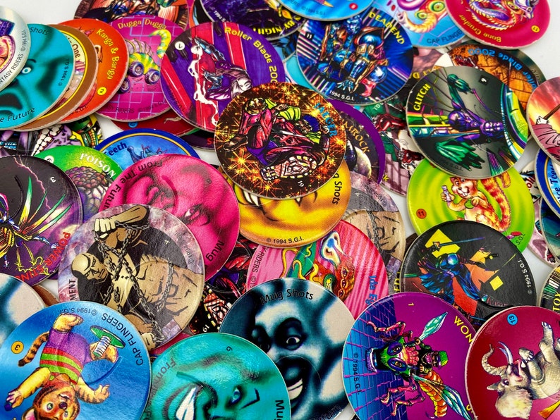 May include: A collection of circular cardboard game discs featuring colourful graphic illustrations of various characters, creatures, and text such as Mug Shots, Cap Flingers, and Glitch. The discs are layered in a pile, displaying diverse artistic styles, vibrant colours, and printed copyright dates from 1994.