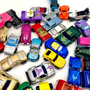 May include: A collection of various miniature toy cars in assorted colours including pink, purple, red, blue, yellow, gold, and green. The vehicles feature diverse designs such as convertibles, coupes, and trucks, scattered across a plain surface.