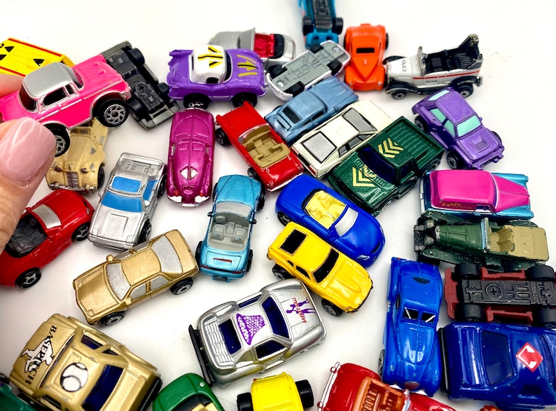 May include: A collection of various miniature toy cars in assorted colours including pink, purple, red, blue, yellow, gold, and green. The vehicles feature diverse designs such as convertibles, coupes, and trucks, scattered across a plain surface.