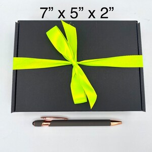 May include: A rectangular black gift box measuring 17.78 cm by 12.7 cm by 5.08 cm, tied with a bright neon yellow ribbon in a bow. Below the box lies a matte dark grey pen with rose gold accents, featuring a clip and a stylus tip.