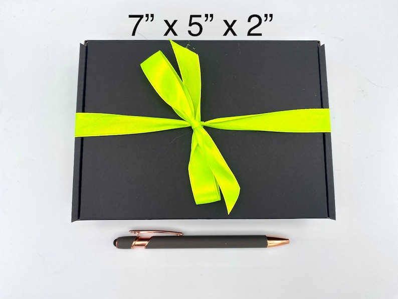 May include: A rectangular black gift box measuring 17.78 cm by 12.7 cm by 5.08 cm, tied with a bright neon yellow ribbon in a bow. Below the box lies a matte dark grey pen with rose gold accents, featuring a clip and a stylus tip.