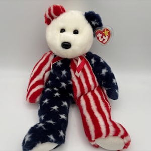 May include: A patriotic plush bear with a white head, red and blue striped arms and legs, and a blue body with white stars. The bear has a red ribbon around its neck and a TY tag.