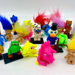 May include: A collection of various small plastic figurines including several troll dolls with vibrant hair, a white Stay Puft Marshmallow Man, a Donald Duck figure, a green Yoshi, a brown E.T. figure, and a pink triceratops head on black stands.