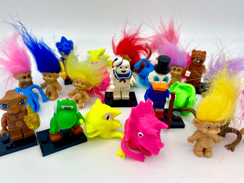 May include: A collection of various small plastic figurines including several troll dolls with vibrant hair, a white Stay Puft Marshmallow Man, a Donald Duck figure, a green Yoshi, a brown E.T. figure, and a pink triceratops head on black stands.