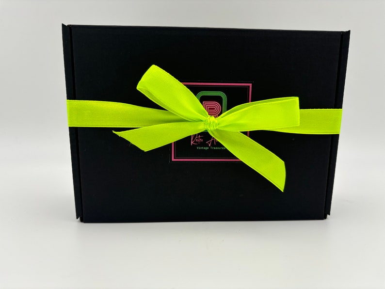 May include: A rectangular black cardboard box wrapped with a bright neon yellow satin ribbon tied in a bow. Centered on the front is a square logo featuring a stylised letter R in green and pink, with the text Retro A Vintage Treasures below.