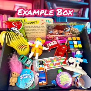 May include: A black box filled with assorted retro toys and sweets, including a yellow spring, a small green car, a troll doll with pink hair, a Rubik's cube, a Smurf figurine, a Donald Duck figure, a dolphin disc, and various snack packages.