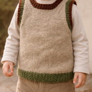 Könnte beinhalten: Ein ärmelloser Strickpullunder in hellem Beige mit dunkelbraunem, geripptem Rundhalsausschnitt und dunkelgrünen Rippbündchen an den Armausschnitten und am unteren Saum, getragen über einem cremefarbenen Langarmshirt und einer beigen Cordhose.