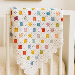 May include: A white crochet blanket featuring a repeating pattern of small, multi-colored squares in shades of red, yellow, blue, green, and pink. The blanket has a delicate, scalloped edge and is draped over the white wooden slats of a crib.